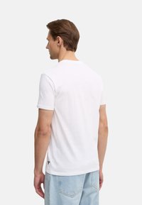 T-shirt bianco in cotone con maniche corte, scollo a girocollo e un piccolo logo all'orlo. Tessuto liscio e forma aderente, indossato con jeans blu chiaro.