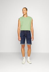 Maglietta senza maniche verde, pantaloni corti di jeans scuri con risvolti, indossati con sneakers alte bianche. Un outfit semplice e informale.