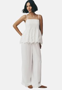 Débardeur blanc avec smocks et bretelles réglables, orné de broderies florales à l'ourlet, associé à un pantalon large blanc ample et des chaussures plates.