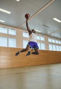 Jugador de baloncesto con una camiseta sin mangas blanca y pantalones cortos morados con acentos verdes, saltando y alcanzando una pelota de baloncesto roja y blanca en un interior.
