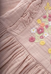 Ružová látka so zhlukovanou textúrou obsahuje farebnú kvetinovú výšivku v žltej, ružovej a bielej farbe. Mäkký, ľahký materiál zo zmesi bavlny.