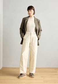 Blazer verde oliva sopra un maglione crema a trama, abbinato a pantaloni beige a vita alta. Scarpe grigie con lacci e suola piatta completano il look.