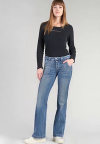 Vaqueros acampanados de mezclilla azul con cinco bolsillos, lavado claro, ligero desgaste y costuras contrastantes, combinados con una camiseta de manga larga negra.