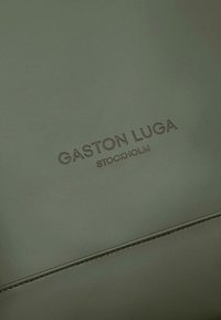 Olive green leather material with embossed "GASTON LUGA STOCKHOLM" branding; smooth texture with subtle stitching detail. Minimalist design.