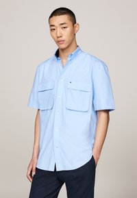Light blue short-sleeve shirt with button-down collar, two front pockets, and small logo on left chest. Fabric appears smooth and lightweight.