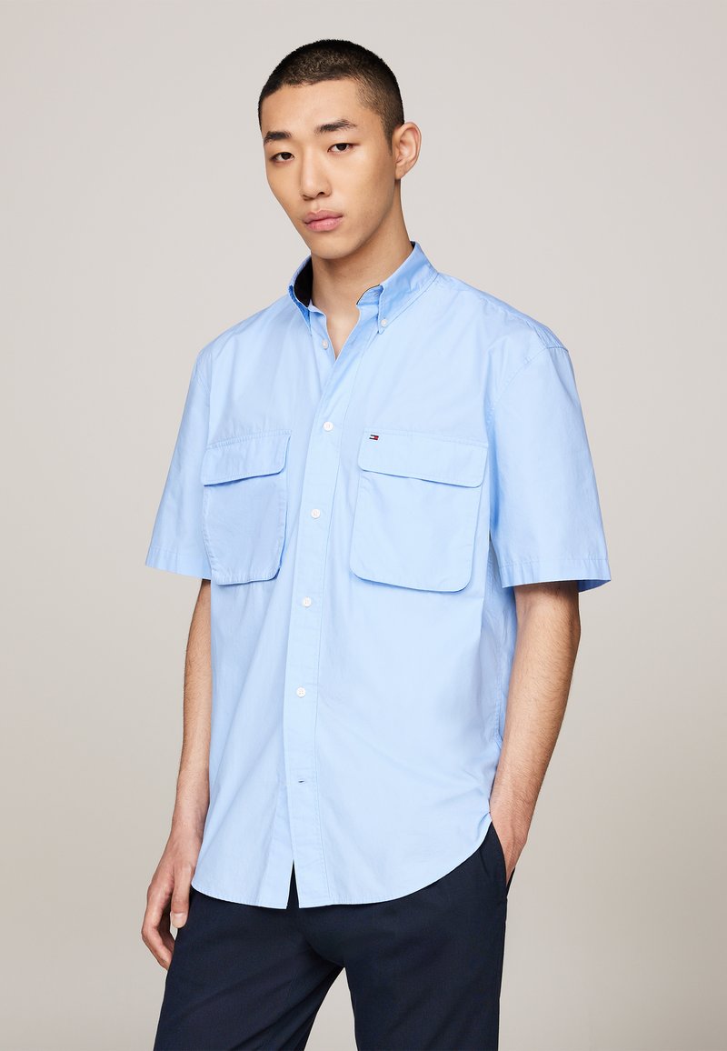 Light blue short-sleeve shirt with button-down collar, two front pockets, and small logo on left chest. Fabric appears smooth and lightweight.