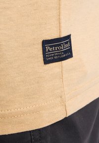 Beige katoen t-shirt met textuurstof, met een marineblauw label waarop "Petrol Ind" staat en stikseldetails langs de zoom.