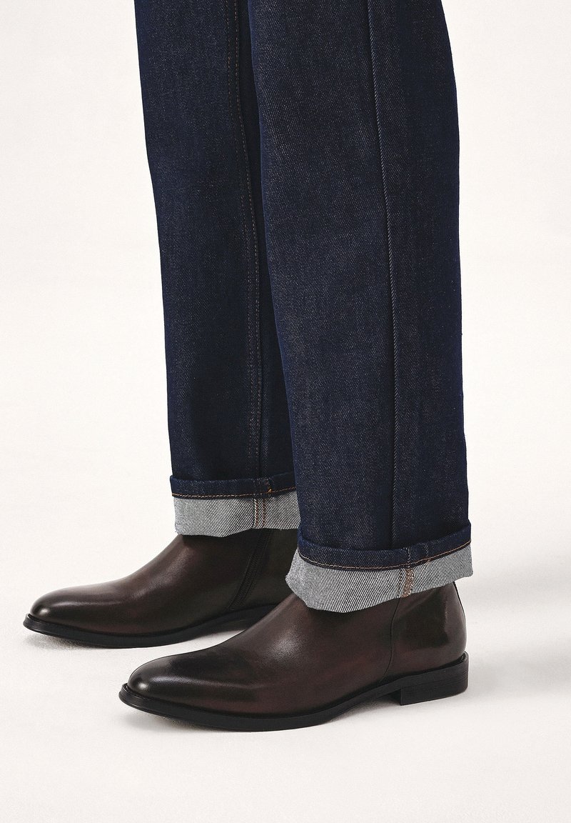 Bottes Chelsea en cuir marron foncé portées avec un jean denim bleu foncé retroussé contre un fond uni clair.