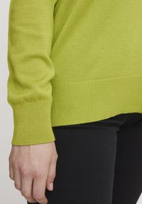 Grüner gestrickter Pullover mit gerippten Bündchen und Saum, der eine weiche Textur und eine entspannte Passform aufweist, kombiniert mit schwarzen, eng anliegenden Hosen.