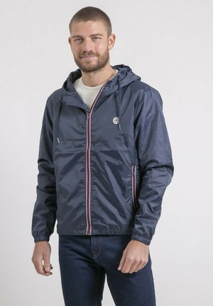 Homme portant un coupe-vent à capuche bleu marine avec une fermeture éclair rayée et des poignets élastiques, associé à un jean bleu foncé, debout devant un fond uni.