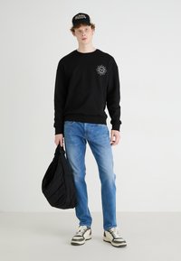 Black sweatshirt with sun graphic, blue jeans, black bag, and sneakers with white and brown accents. Casual outfit on neutral background.