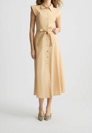 Femme portant une robe midi beige sans manches boutonnée avec une ceinture à la taille et des talons pointus à motifs, debout devant un fond uni.