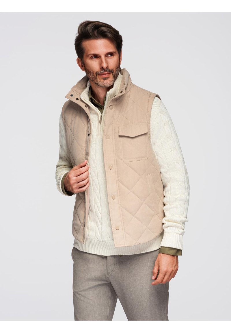 Gilet imbottito beige con colletto alto, tasche frontali e chiusure a scatto, indossato sopra un maglione a maglia a trecce di colore crema.