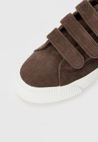 Brown corduroy sneaker with three adjustable Velcro straps and a white rubber sole. Features ridged texture and rounded toe design.