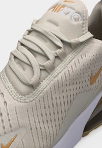 Baskets Nike Air 270 beige clair avec tissu texturé, languette rembourrée, lacets beiges et logos Nike swoosh orange sur le côté et la pointe.