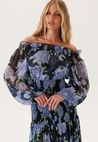 Vrouw in een off-shoulder jurk met doorzichtige geplooide mouwen en een blauwe bloemenprint, met haar handen zachtjes samengevouwd op de taille.