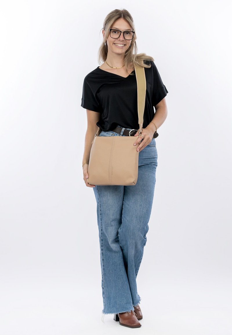 Borsa a tracolla in pelle beige con forma rettangolare piatta, dettagli di cucitura visibili e tracolla regolabile. Indossata da una persona in abbigliamento casual.
