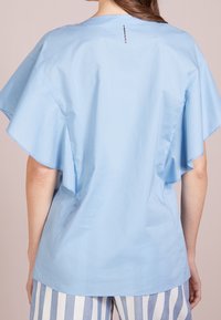Blusa azzurra chiara con maniche corte e larghe e una vestibilità rilassata, realizzata in un tessuto leggero e morbido. Presenta un dettaglio di cucitura sul retro.