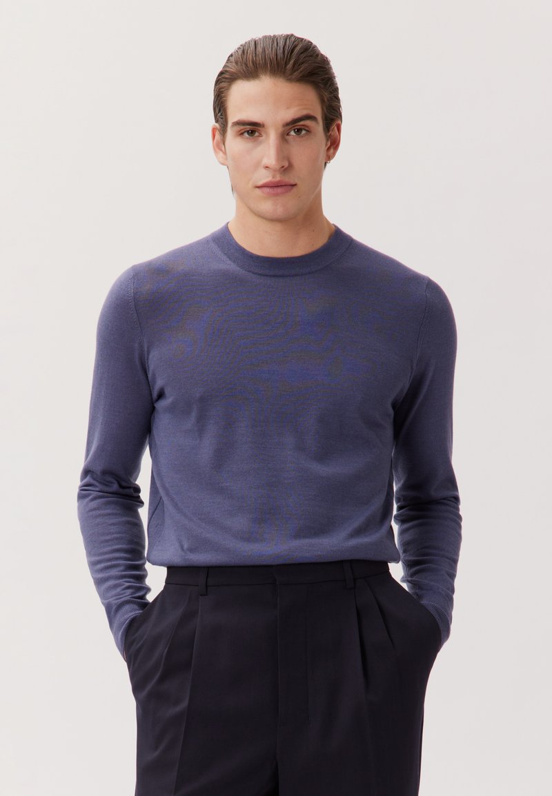 Jeune homme aux cheveux lissés en arrière, portant un pull violet à col rond et un pantalon noir plissé, mains dans les poches, fond neutre.