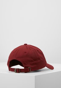 Cappellino da baseball in cotone rosso con visiera curva, straps regolabile e fori per la traspirazione. Mostra la vista posteriore su uno sfondo neutro.
