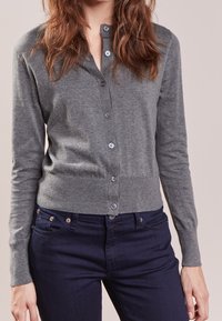 Harmaa neuletakki, jossa on napitus, pitkät hihat ja istuva malli. Päällä tummansiniset farkut, joissa on hienovarainen resorikuvio.