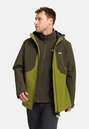 Mann trägt zweifarbige grüne Outdoorjacke und dunkle Hose, schaut nach links mit entspannt an den Seiten hängenden Händen vor einfarbigem Hintergrund.