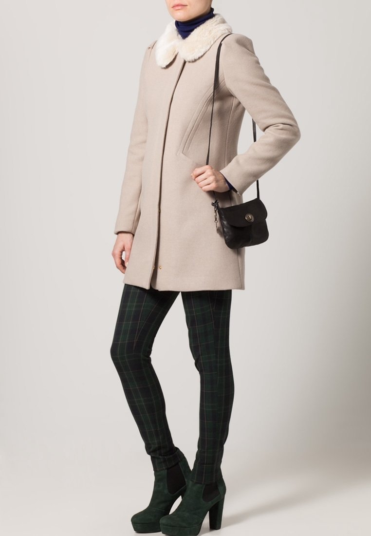 Personne portant un manteau beige avec col en fausse fourrure blanche, un sac bandoulière noir, des leggings à carreaux foncés, et des bottines vertes à talons hauts.