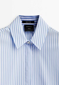 Chemise à rayures verticales bleu et blanc à boutons, avec col pointu et étiquettes noires à l'intérieur du col.