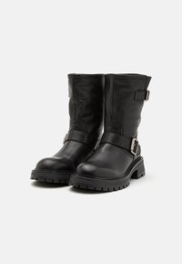 Botas de couro pretas até ao joelho, com bico arredondado, tiras laterais e fivelas metálicas. A sola é texturizada para agarrar.