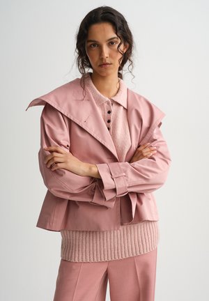 Kobieta o ciemnych kręconych włosach ubrana w różowy sweter z kołnierzykiem, luźną różową kurtkę i dopasowane spodnie, stojąca z założonymi rękami na jasnym tle.