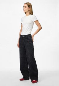 Naine seisab käsi puusal, seljas slim-fit valge särk, laiad mustad teksad ja punased kingad, halli valge tausta ees.
