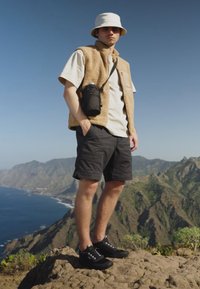 Beige fleece vest over a light shirt, black shorts, and black shoes. Accessories include a black camera bag and a white bucket hat. Mountain backdrop.