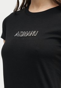 T-shirt noir à col rond avec le logo « ARMANI EXCHANGE » en lettres blanches et noires texturées sur la poitrine, porté par une personne aux cheveux longs.