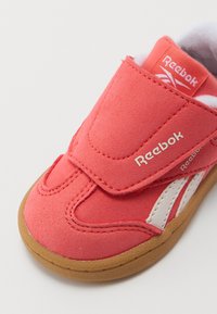 Chaussure Reebok corail avec une sangle velcro, présentant une tige semblable à du daim, des accents blancs et une semelle en caoutchouc brun.