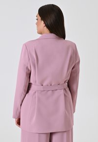 Lichtroze op maat gemaakte blazer met een ceintuur in de taille, schoudervullingen en een gladde, satijnen uitstraling. De achterzijde toont strakke lijnen en naaddetails.