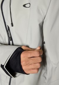 Pilkas vandeniui atsparus švarkas su juodu vidiniu rankogaliu, reguliuojamu virvute ir užtrauktukais rankovėje, užtikrinančiais tvirtą prigludimą.