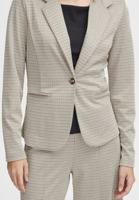 Veste de costume à carreaux beige avec une fermeture à un bouton, coupe ajustée et deux poches avant, portée avec un haut noir en dessous.