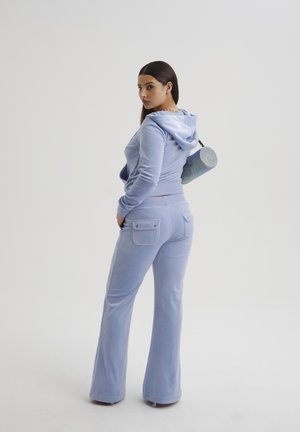 Tuta in velluto blu con hoodie corta e pantaloni svasati dotati di tasche laterali; modello che tiene in mano una borsa cilindrica scintillante.