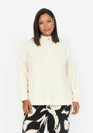 Sudadera de color crema con cuello alto y cierre de media cremallera. Tejido suave y liso con mangas largas y un ajuste relajado. Combinada con pantalones estampados.