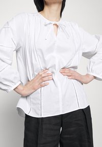 Blusa bianca con scollatura rotonda, dettaglio con fiocco nella parte anteriore e cuciture decorative lungo i lati. Maniche lunghe e puffate con taglio rilassato.