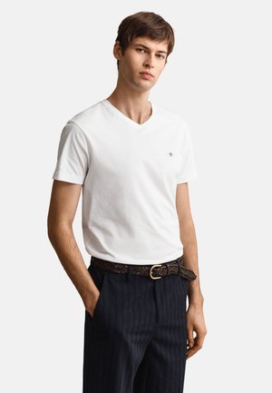 Jeune homme portant un T-shirt blanc col en V rentré dans un pantalon foncé à fines rayures avec une ceinture tressée marron, les mains dans les poches.