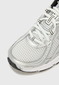 Chaussure de sport argentée avec une tige en mesh, lacets blancs, semelle en caoutchouc noire et détails texturés sur les côtés. Fini légèrement réfléchissant.