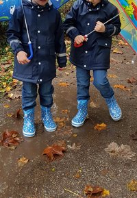 Bottes de pluie en caoutchouc bleues avec des motifs floraux colorés,portées par des enfants en manteaux foncés, se tenant dans une flaque sur un chemin jonché de feuilles.