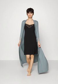 Črna obleka brez rokavov z čipkasto obrobo, združena z dolgim svetlo sivim jopico z vzorčastimi rokavi. Model je bosa v nevtralnem okolju.