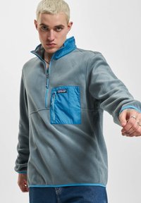 Pullover in pile grigio con colletto a mezza zip, dotato di tasca sul petto turchese e orlo a contrasto. Texture liscia con dettagli di cucitura visibili.