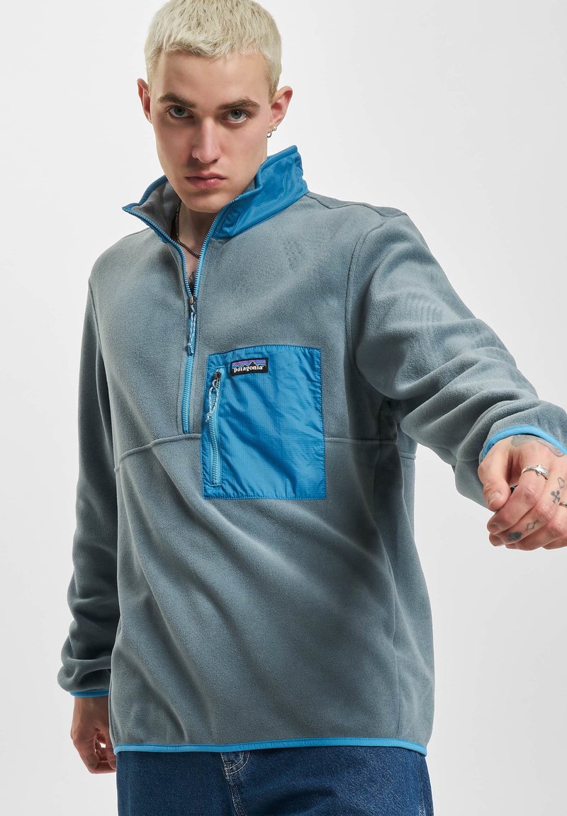 Pullover in pile grigio con colletto a mezza zip, dotato di tasca sul petto turchese e orlo a contrasto. Texture liscia con dettagli di cucitura visibili.
