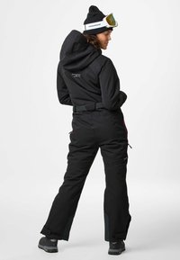 Black ski overall with a hood, adjustable belt, two zippered pockets, and contrasting pink accents. Designed for winter sports, durable fabric.