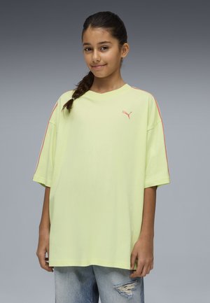 Meisje met gevlochten haar, gekleed in een oversized lichtgeel Puma-shirt en gescheurde blauwe spijkerbroek, staand tegen een grijze verloopachtergrond.