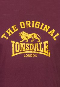 Tricou din bumbac maron cu un imprimeu de text galben, uzat, care spune "THE ORIGINAL LONSDALE LONDON" și cu un grafic cu un leu.