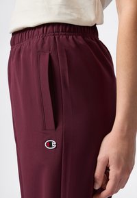 Close-up van een persoon die bordeauxrode broek draagt met een elastische tailleband, zijzak en een klein Champion-logo dicht bij de zak.
